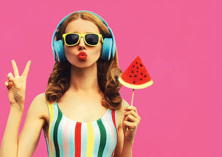 Summer portrait of stylish woman in headphones listening to music blowing her lips with juicy lollipop or ice cream shaped slice watermelon on pink background, blank copy space for advertising text