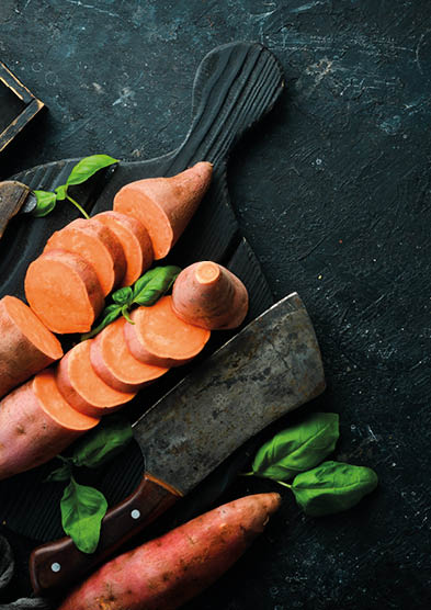 Raw sweet potatoes and basil on a black stone background. Top view. Free space for your text.