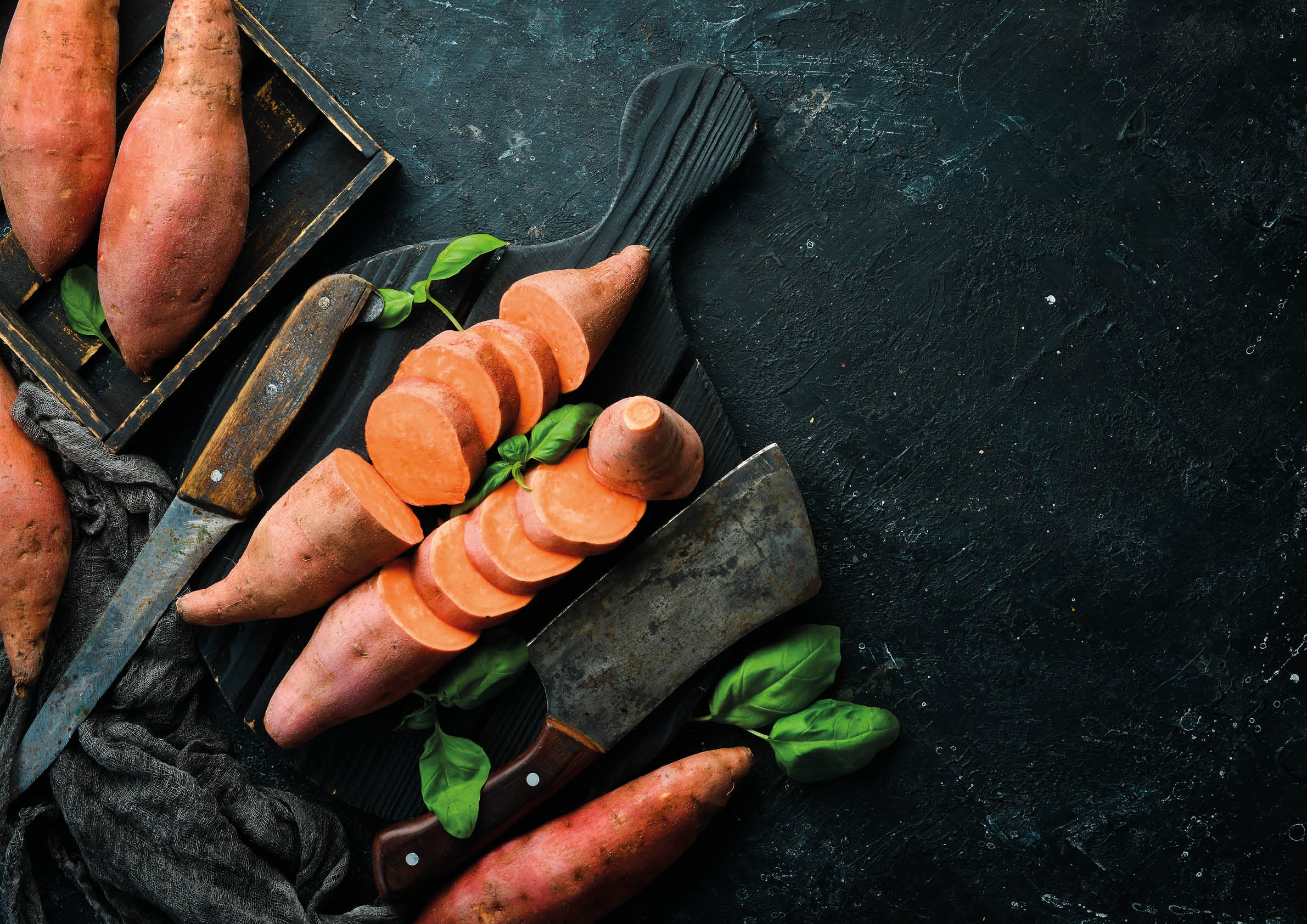 Raw sweet potatoes and basil on a black stone background. Top view. Free space for your text.